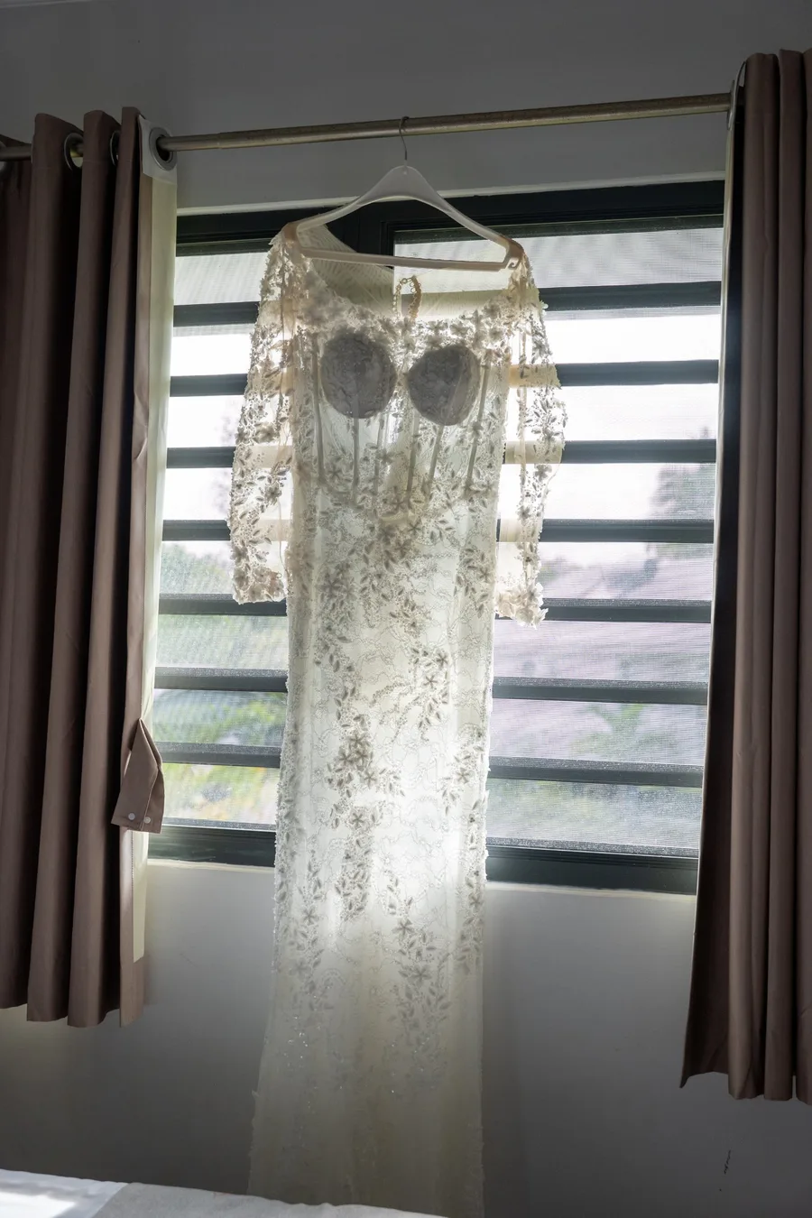 Robe de mariée en dentelle perlée suspendue devant une fenêtre